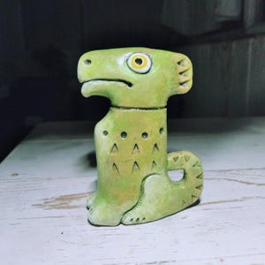 Pode incluir: Uma estatueta de cerâmica verde-clara de uma criatura estilizada com uma cabeça grande, um focinho proeminente semelhante a um bico e um olho amarelo visível com pupila preta. O corpo apresenta padrões triangulares incisos e uma cauda.