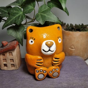 May include: A small, orange ceramic bear figurine with a white muzzle, black eyes, and grey paw pads. The bear is depicted in a seated position with its arms resting on its knees. The surface of the ceramic has a glossy finish and rounded features.