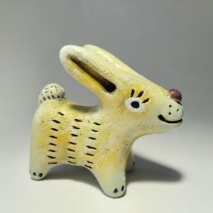 Może przedstawiać: Ręcznie malowana ceramiczna figurka królika w odcieniach żółci i kremu. Królik ma czarne oznaczenia, mały, okrągły ogon i czerwony nos. Jego oczy są obrysowane na biało i ma uśmiechniętą minę. Przedmiot dekoracyjny.