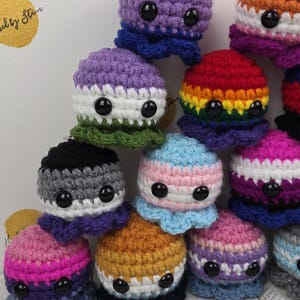 May include: A collection of colorful, crocheted octopus plushies. Each octopus features a different color scheme, including rainbow, pink, purple, and blue, with black button eyes. The background is a light color with the text "Hooked by Star".