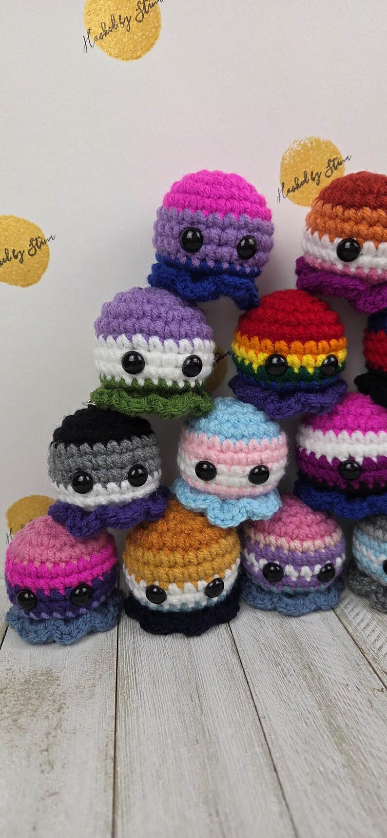 May include: A collection of colorful, crocheted octopus plushies. Each octopus features a different color scheme, including rainbow, pink, purple, and blue, with black button eyes. The background is a light color with the text "Hooked by Star".