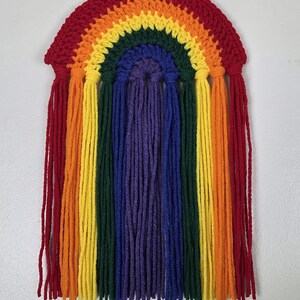 May include: A wall hanging featuring a crocheted rainbow arch in red, orange, yellow, green, blue, and purple. Long, vertical yarn fringe in matching colors hangs from the bottom of each arch section, creating a layered, textured appearance.