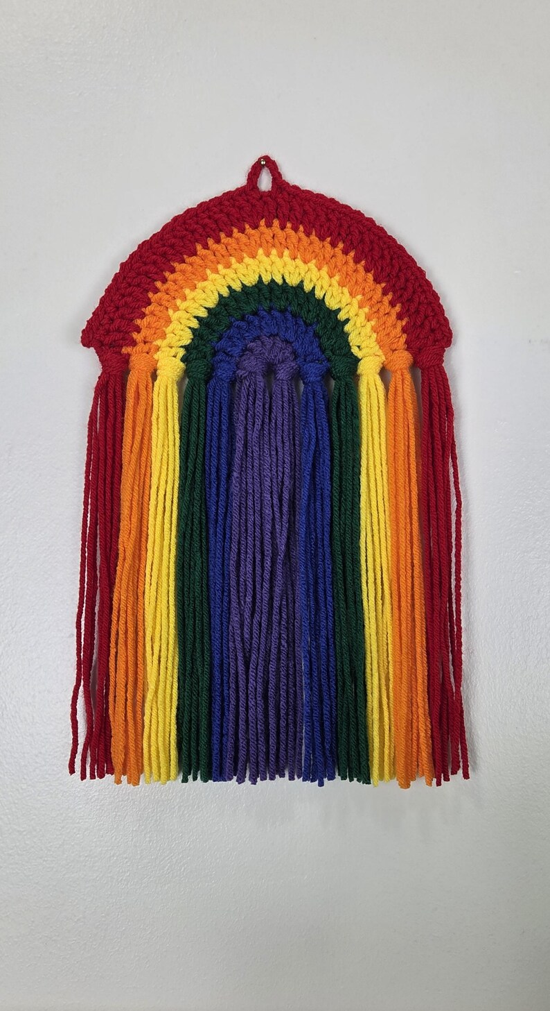 May include: A wall hanging featuring a crocheted rainbow arch in red, orange, yellow, green, blue, and purple. Long, vertical yarn fringe in matching colors hangs from the bottom of each arch section, creating a layered, textured appearance.