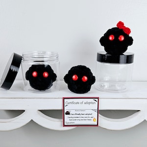 May include: Three black crocheted demon plushies with red eyes. Two are in clear jars with black lids. The third is on a white shelf with a red bow. A red and black certificate of adoption is on the shelf. The certificate reads "Has officially been adopted! I hereby promise to love, care for, and feed souls to my new best friend. Signature:"