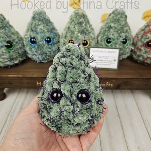 May include: A green crocheted plush toy shaped like a cannabis plant with purple accents and large black eyes. The toy is being held in a person's hand.