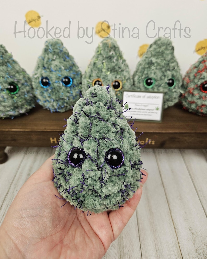 May include: A green crocheted plush toy shaped like a cannabis plant with purple accents and large black eyes. The toy is being held in a person's hand.