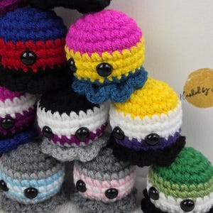 May include: A collection of handmade crochet octopus plushies in various colors, each representing different LGBTQ+ pride flags. The plushies have black button eyes and are stacked on a wooden surface. The text "Hooked by Stina" is visible.