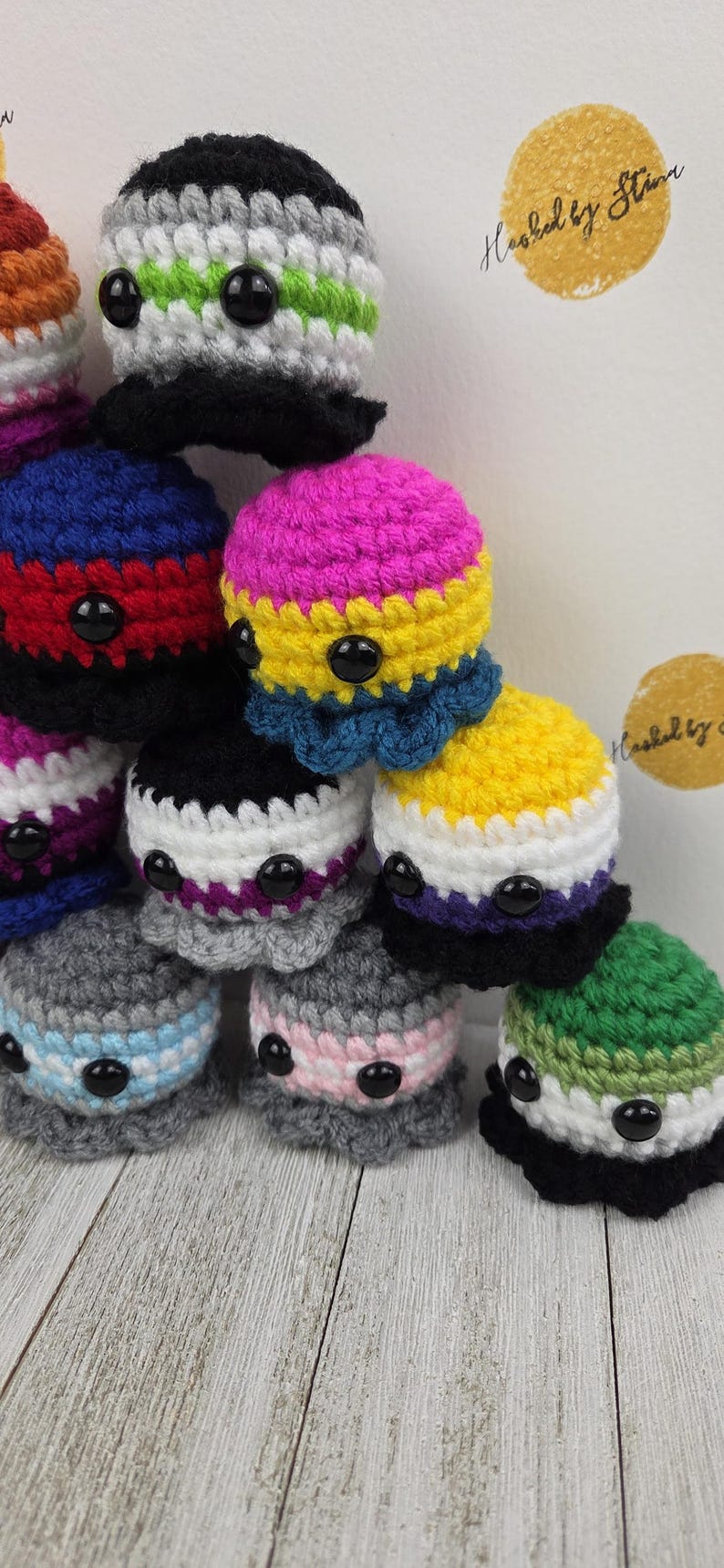 May include: A collection of handmade crochet octopus plushies in various colors, each representing different LGBTQ+ pride flags. The plushies have black button eyes and are stacked on a wooden surface. The text "Hooked by Stina" is visible.
