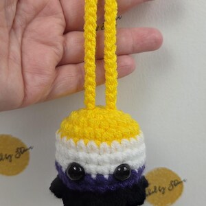 May include: A handmade crochet ornament in the non-binary pride flag colors: yellow, white, purple, and black. The ornament has a yellow top with a loop for hanging, and two black bead eyes.
