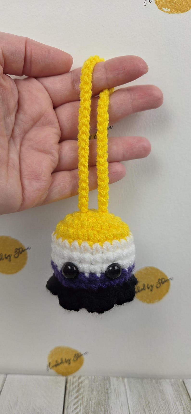 May include: A handmade crochet ornament in the non-binary pride flag colors: yellow, white, purple, and black. The ornament has a yellow top with a loop for hanging, and two black bead eyes.