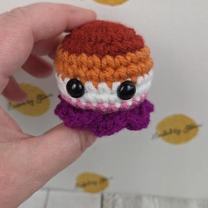 May include: A handmade crochet octopus in the colors of the lesbian pride flag: dark orange, orange, white, pink, and purple. The octopus has large black eyes and a ruffled skirt. The text "Hooked by Star" is visible in the background.