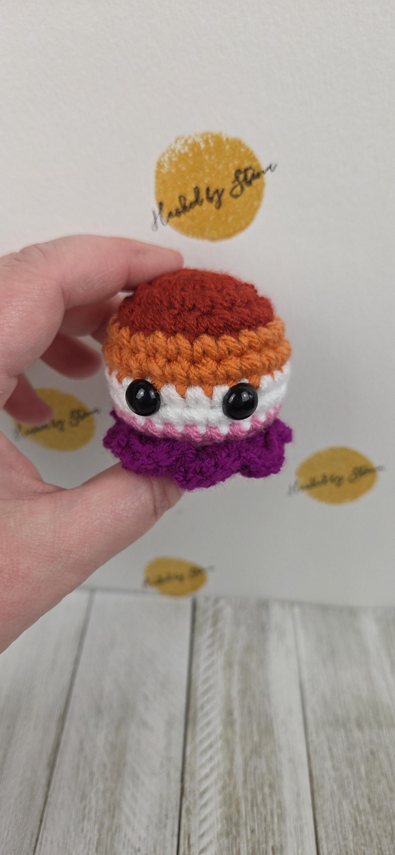 May include: A handmade crochet octopus in the colors of the lesbian pride flag: dark orange, orange, white, pink, and purple. The octopus has large black eyes and a ruffled skirt. The text "Hooked by Star" is visible in the background.