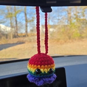 May include: A handmade crochet octopus car charm in vibrant rainbow colors. The octopus has black bead eyes and a red top, with a rainbow body and purple tentacles. The charm hangs from a red cord.