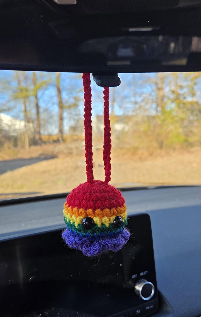 May include: A handmade crochet octopus car charm in vibrant rainbow colors. The octopus has black bead eyes and a red top, with a rainbow body and purple tentacles. The charm hangs from a red cord.