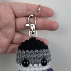 May include: Crocheted keychain featuring an asexual pride flag design. The keychain has a black top, gray middle, white bottom, and purple frill. It includes a silver-colored clasp and ring.
