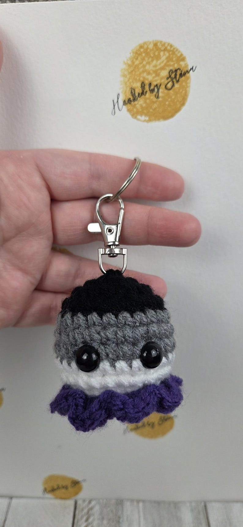 May include: Crocheted keychain featuring an asexual pride flag design. The keychain has a black top, gray middle, white bottom, and purple frill. It includes a silver-colored clasp and ring.