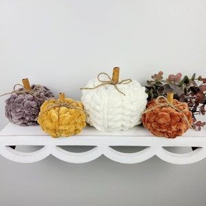 May include: Four yarn pumpkins in different colors: gray, mustard yellow, white, and rust orange. The pumpkins are arranged on a white shelf with a scalloped edge.