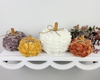 May include: Four yarn pumpkins in different colours: grey, mustard yellow, white and rust orange. The pumpkins are arranged on a white shelf with a scalloped edge.