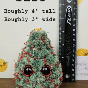 May include: A green and red crocheted Christmas tree plush toy with googly eyes. The toy is approximately 4 inches tall and 3 inches wide. The toy is sitting on a wooden surface.