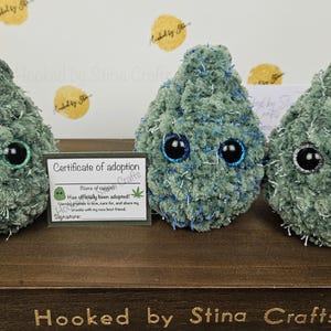 May include: Three green plush toys shaped like marijuana buds with googly eyes. The toys are sitting on a wooden surface with a certificate of adoption card in front of them. The card reads "Certificate of adoption" and "Has officially been adopted!"