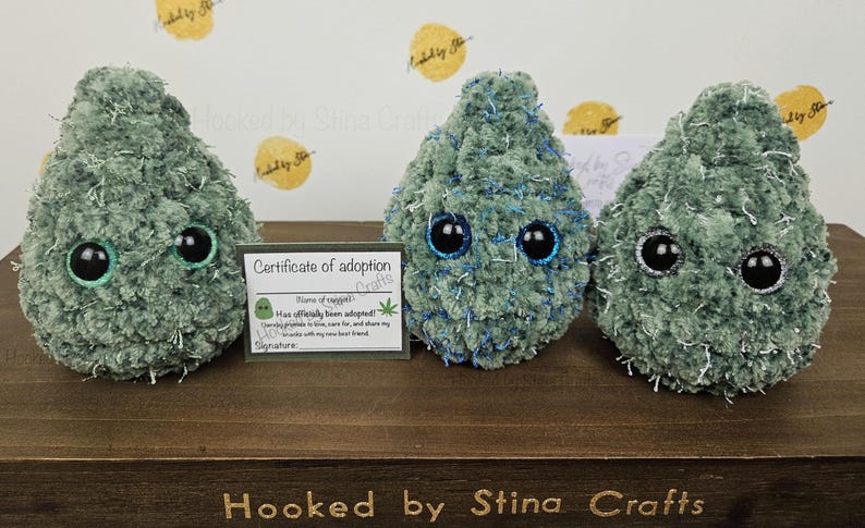 May include: Three green plush toys shaped like marijuana buds with googly eyes. The toys are sitting on a wooden surface with a certificate of adoption card in front of them. The card reads "Certificate of adoption" and "Has officially been adopted!"