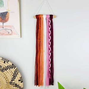 May include: A wall hanging made of yarn in shades of burgundy, orange, white, and pink. The yarn is woven into a variety of patterns, including braids and knots. The hanging is attached to a wooden dowel.
