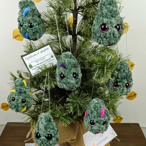 May include: A small artificial Christmas tree decorated with handmade crocheted ornaments shaped like marijuana buds. The ornaments have googly eyes and are green with various colored accents. The tree is in a burlap pot and there is a sign that says "Hooked by Stina Crafts".
