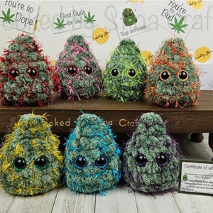 May include: A collection of handmade, plush, bud-shaped toys in various colors, including green, red, pink, and blue. Each toy has large, googly eyes and a fluffy texture. The toys are displayed on a wooden shelf, with small signs that read "You're so Dope" and "Best Buds for life!"