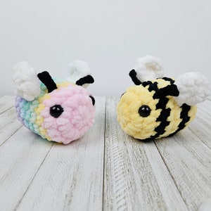 May include: Two crocheted bee plushies. One bee is pink, yellow, and blue with white wings. The other bee is yellow and black with white wings.