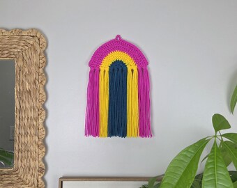 May include: A wall hanging featuring a crocheted rainbow arch in magenta, yellow, and dark blue. Long, straight yarn tassels in the same three colors hang vertically from the bottom of the arch, creating a layered, textured fiber art piece.