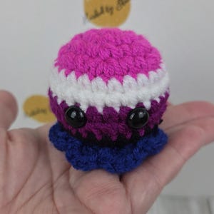 May include: A handmade crochet octopus in shades of pink, white, purple, and blue. The octopus has large black eyes and a ruffled base. The toy is held in a hand.
