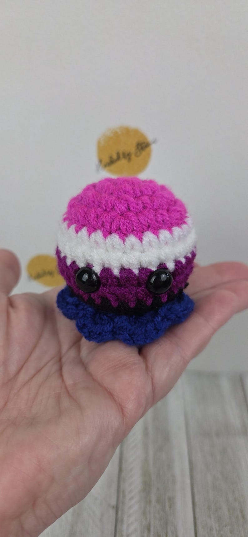 May include: A handmade crochet octopus in shades of pink, white, purple, and blue. The octopus has large black eyes and a ruffled base. The toy is held in a hand.