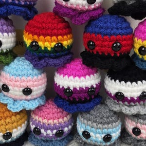 May include: A collection of handmade crochet octopus plushies in various colors, each representing different LGBTQ+ pride flags. The plushies have black button eyes and are arranged on a light wooden surface.