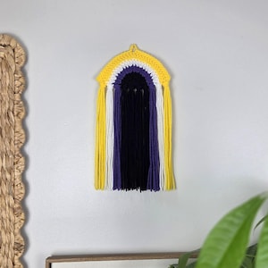 May include: A wall hanging featuring a crocheted arch in yellow, white, and purple, with long, straight yarn fringe hanging down in vertical stripes of yellow, white, purple, and black, creating a layered, textured, and colorful fiber art piece.