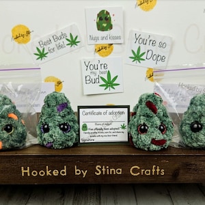 May include: Four green plush weed bud toys with googly eyes and different facial expressions. The toys are packaged in clear plastic bags with purple zippers. The bags are sitting on a wooden surface with the text "Hooked by Stina Crafts" printed on it. There are several small cards with various messages and images of weed buds.