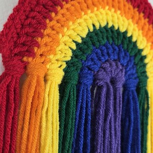 May include: A semicircular wall hanging crafted from crocheted yarn in a rainbow pattern. The design features concentric arcs of red, orange, yellow, green, blue, and purple, with long, loose yarn tassels hanging vertically from the bottom edge.