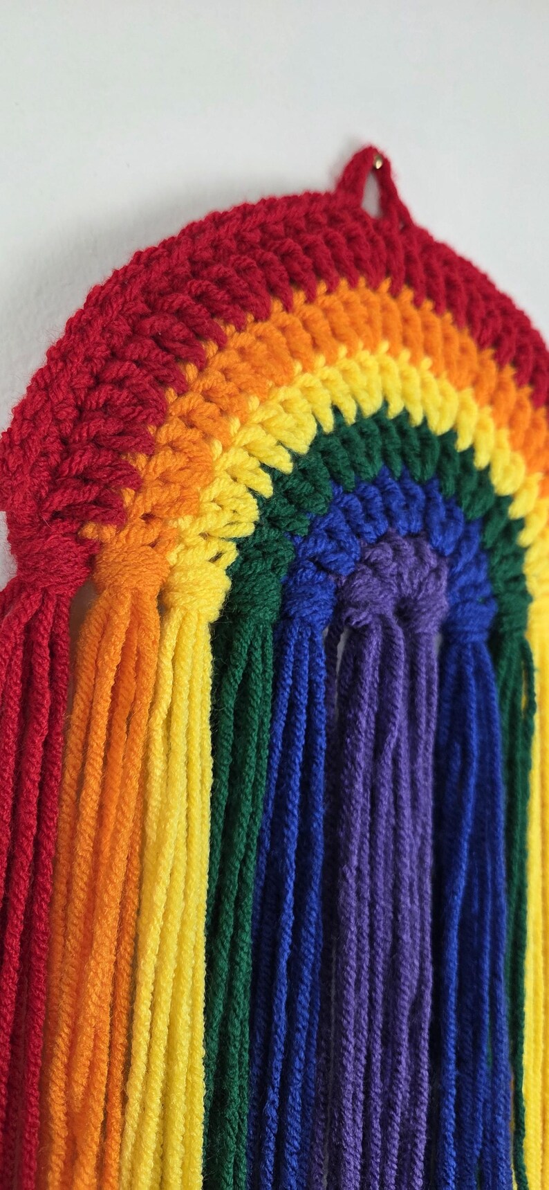 May include: A semicircular wall hanging crafted from crocheted yarn in a rainbow pattern. The design features concentric arcs of red, orange, yellow, green, blue, and purple, with long, loose yarn tassels hanging vertically from the bottom edge.
