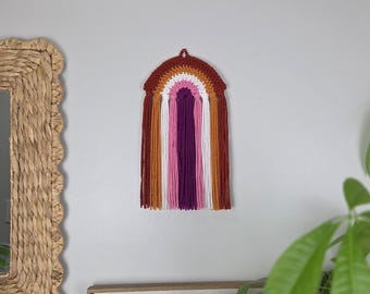 May include: A wall hanging featuring a crocheted rainbow arch in shades of dark red, orange, white, pink, and purple. Long, vertical yarn fringe in matching colors hangs from the bottom of the arch, creating a textured, layered appearance.
