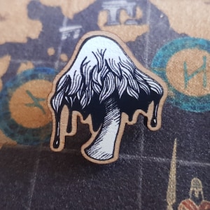 May include: A wooden pin featuring a black and white illustration of a mushroom with dripping black paint. The mushroom has a white cap with black lines and a white stem.