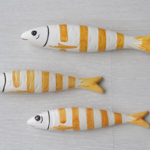 Op de afbeelding: Drie houten visfiguren met witte en gele strepen. De vissen zijn geschilderd met zwarte ogen en een eenvoudige glimlach.