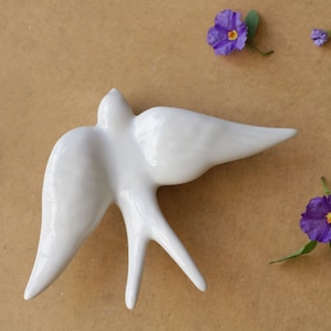 May include: White ceramic bird figurine with a stylized design, resembling a swallow in flight. The figurine is on a brown background with purple flowers.
