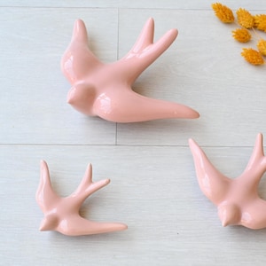 Pode incluir: Três andorinhas de cerâmica rosa em voo, dispostas sobre uma superfície clara com grão de madeira. As aves têm as asas abertas e são de vários tamanhos. Flores amarelas secas no canto superior direito.