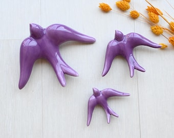May include: Three glossy, purple ceramic swallows in varying sizes are arranged on a light-colored surface. The birds have outstretched wings and detailed tail feathers. Dried yellow flowers are in the background.