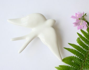 May include: White ceramic swallow bird wall decor. The bird is in flight with its wings spread. The bird is a simple design with no details.