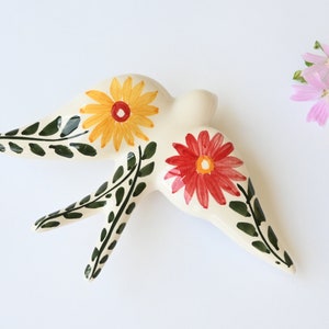 May include: A ceramic swallow figurine, painted with yellow and red flowers and green leaf accents. The bird is cream-colored and has a stylized design. A small bouquet of pink and yellow flowers is to the right.
