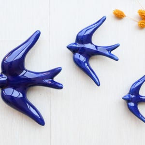 Puede incluir: Tres golondrinas de cerámica azul cobalto brillante de diferentes tamaños están dispuestas sobre una superficie blanca. Las aves tienen las alas extendidas y están posicionadas como si estuvieran en vuelo. Flores naranjas secas en la esquina superior derecha.
