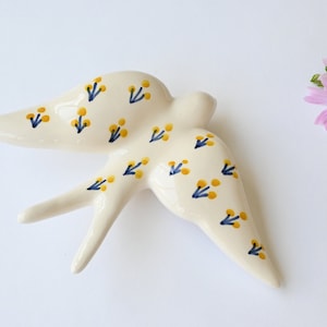 Könnte beinhalten: Cremefarbene Keramik-Schwalbenfigur mit gelben und marineblauen Blumenmustern. Der Vogel ist im Flug mit ausgebreiteten Flügeln dargestellt. Rechts ein kleiner Strauß rosa und gelber Blumen. Ein dekorativer Artikel.
