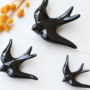 Può includere: Tre rondini in ceramica nera lucida di varie dimensioni sono disposte su una superficie bianca, con un rametto di fiori arancioni secchi. Gli uccelli hanno le ali spiegate, suggerendo il volo.