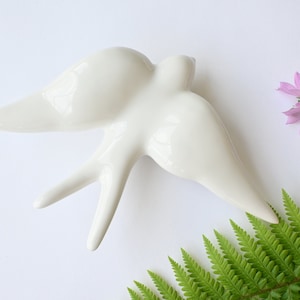 May include: White ceramic swallow bird wall decor. The bird is in flight with its wings spread out. The bird is a simple design with no details.