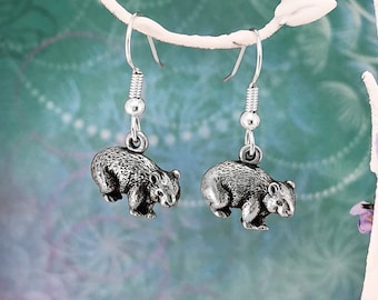 May include: A pair of silver dangle earrings featuring a detailed 3D wombat charm. The earrings are hanging from a white display stand.